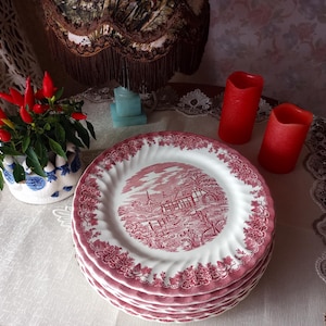 Può includere: Una pila di piatti piani in ceramica bianca con bordo smerlato e un motivo transferware rosso scuro. Il design centrale raffigura un paesaggio rurale con edifici e alberi, mentre il bordo è decorato con una dettagliata bordura floreale.