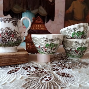 Könnte beinhalten: Eine weiße Keramik-Sahnekännchen mit einer braunen und schwarzen Landschaftsszene, ein Stapel Untersetzer und drei Teetassen mit einem ähnlichen Design. Die Teetassen und das Sahnekännchen stehen auf einer weißen Spitzendecke. Im Hintergrund befindet sich ein dekoratives Holzhaus.
