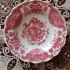 Può includere: Una ciotola rotonda in ceramica con bordo smerlato, caratterizzata da una base bianca e un dettagliato motivo floreale rosa. Il design include un bouquet di fiori centrale e bordi ornamentali a volute lungo il bordo della ciotola.