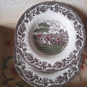 May include: Two white ceramic bowls with a scalloped edge and a dark brown border featuring an acorn and oak leaf pattern. The center of the top bowl displays a colorful scene of people on horseback riding through a field with trees and buildings.