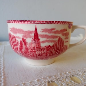 Op de afbeelding: Een crèmekleurige keramische theekop met een rood transferpatroon van een kerk met een hoge toren omringd door bomen. De rand van de kop is versierd met een herhalende geometrische rand en de kop is voorzien van een enkel gebogen handvat.