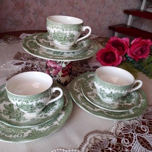 Może przedstawiać: Zestaw trzech białych ceramicznych filiżanek do herbaty z pasującymi spodkami, ozdobionych szczegółowym zielonym wzorem kwiatowym i plecionym obrzeżem. Zestaw zawiera trzy większe talerze, z jedną filiżanką i spodkiem umieszczonymi na cylindrze.