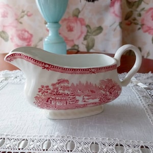 Könnte beinhalten: Eine weiße Keramik-Sauciere mit einem rot-weißen Landschaftsdesign. Das Boot hat einen Griff und einen dekorativen roten Rand. Die Sauciere steht auf einer weißen Spitzendecke.
