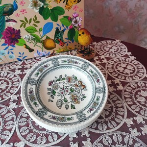 Könnte beinhalten: Ein Stapel weißer Keramikteller mit floralem Design. Die Teller haben einen detaillierten Rand mit einem blau-grauen Muster. Die Mitte der Teller zeigt ein farbenfrohes Blumenarrangement mit Vögeln.