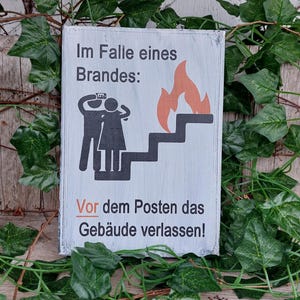 Könnte beinhalten: Ein weißes Holzschild mit schwarzem Text und Illustrationen. Das Schild lautet "Im Falle eines Brandes: Vor dem Posten das Gebäude verlassen!"  Die Illustration zeigt zwei Strichmännchen, die eine Treppe hinuntergehen, über ihnen ein Feuer.
