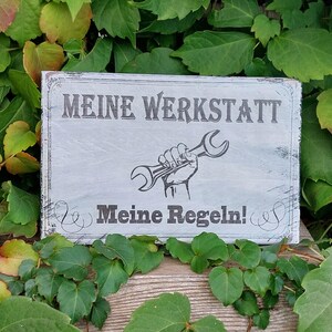 Könnte beinhalten: Ein graues Holzschild mit dem Text "Meine Werkstatt Meine Regeln!" und einer schwarz-weißen Illustration einer Hand, die einen Schraubenschlüssel hält.