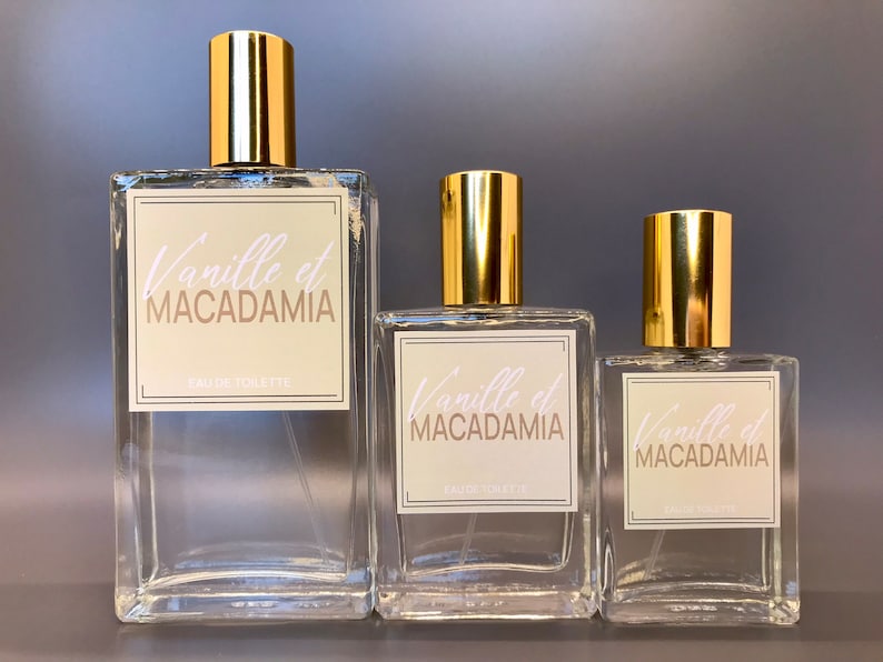 Three glass bottles of perfume, 100ml, 50ml and 30ml with gold caps and labels that say "Vanille et Macadamia" in white script. The bottles are different sizes.