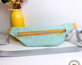 May include: A light teal fanny pack with a yellow zipper and trim. The bag has a gray strap and silver hardware. A yellow vase with flowers is in the background.