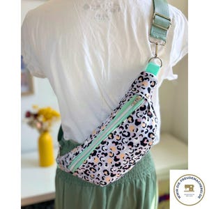 Könnte beinhalten: Eine Bauchtasche mit Leopardenmuster in Schwarz, Pfirsich und Weiß. Der Reißverschluss und der Gurt sind mintgrün. Der verstellbare Gurt wird mit einem silbernen Verschluss befestigt. Die Tasche wird quer über dem Körper getragen.