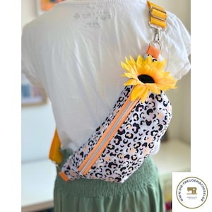 Puede incluir: Riñonera con estampado de leopardo blanco y negro con cremallera y correa naranja. La bolsa está adornada con un girasol amarillo y tiene una correa amarilla. La bolsa se lleva sobre el hombro.