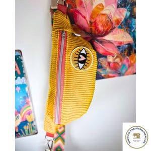 Könnte beinhalten: Senfgelbe Cord-Bauchtasche mit rosa Reißverschluss und einem Augen-Patch. Die Tasche hat einen bunten Gurt und einen runden Aufnäher mit einem Augendesign. Die Tasche hängt an einer weißen Wand.