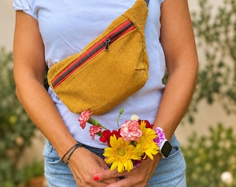 May include: A mustard yellow corduroy fanny pack with a red zipper accent and a patterned, multi-colored woven strap. The bag is worn across the chest over a plain white t-shirt, which features a small embroidered beer bottle graphic on the chest.