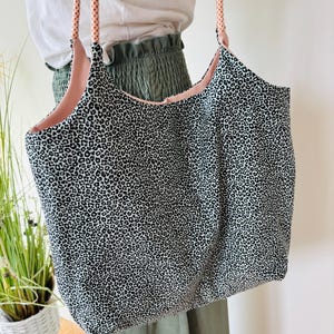 Könnte beinhalten: Eine große Tragetasche mit einem schwarz-weißen Leopardenmuster außen und einem rosa Innenfutter. Die Tasche hat zwei Seilgriffe mit einem pfirsichfarbenen und weißen Tupfenmuster. Die Tasche wird von einer Person gehalten, die ein weißes Oberteil und grüne Hosen trägt.