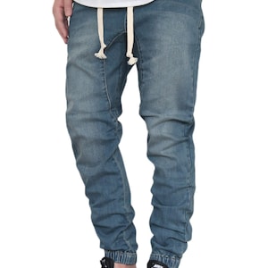 May include: A pair of blue denim jogger pants with a drawstring waist and a cuffed hem. The pants are being worn with black sneakers.