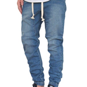 May include: A pair of blue denim jogger pants with a drawstring waist and a cuffed hem. The pants are worn by a person wearing a white t-shirt and black sneakers. The word "INDIGO" is printed in red at the bottom of the image.