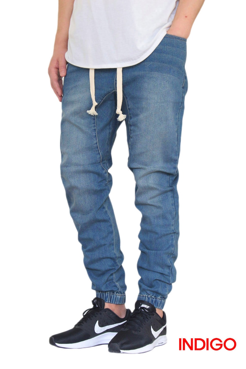 May include: A pair of blue denim jogger pants with a drawstring waist and a cuffed hem. The pants are worn by a person wearing a white t-shirt and black sneakers. The word "INDIGO" is printed in red at the bottom of the image.