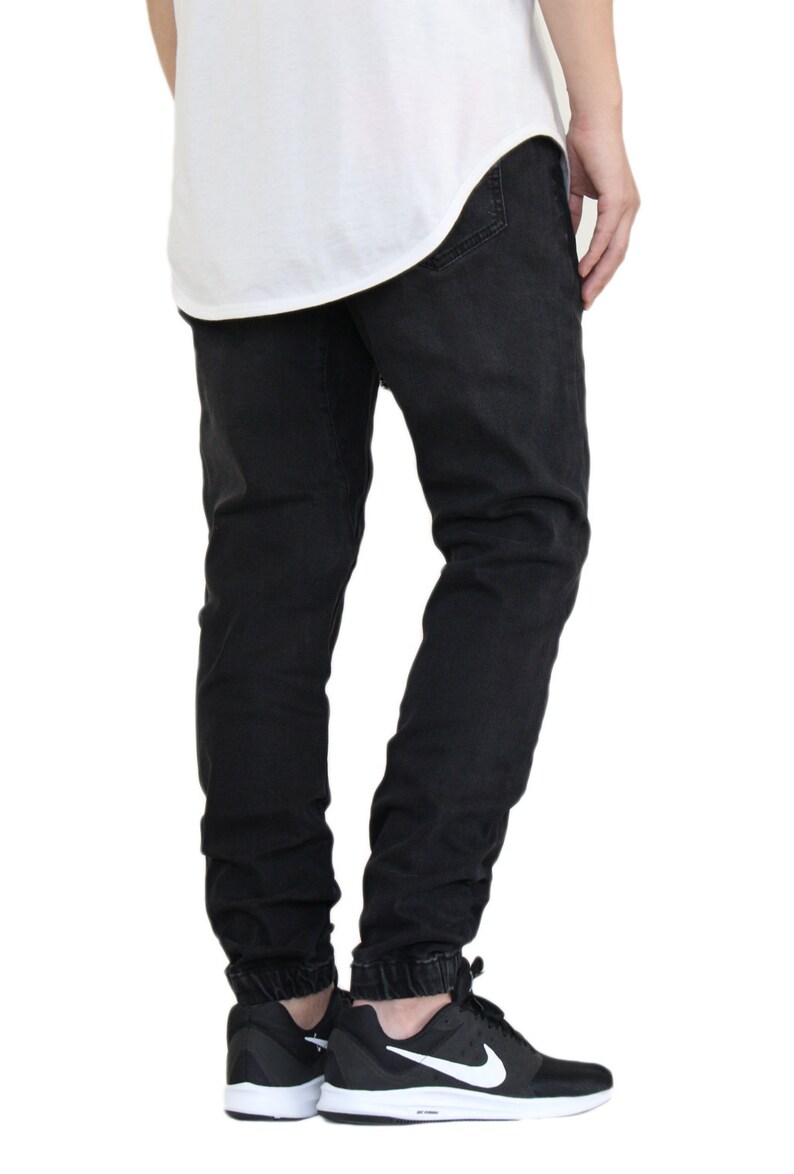 May include: Black jogger style jeans with a cuffed hem. The jeans are worn by a person wearing black and white sneakers.