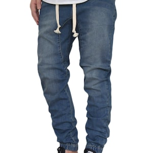 May include: A pair of dark indigo blue denim jogger pants with a drawstring waist and cuffed ankles. The pants are being modeled by a person wearing black sneakers.