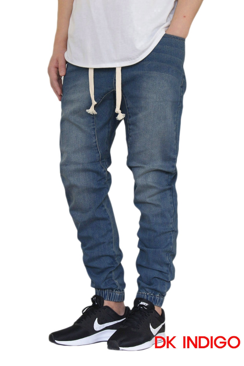 May include: A pair of dark indigo blue denim jogger pants with a drawstring waist and cuffed ankles. The pants are being modeled by a person wearing black sneakers.