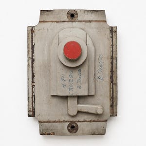 Puede incluir: Un molde industrial rectangular de madera gris con un elemento circular central pintado de rojo, una pequeña palanca y texto escrito a mano que dice H.P.I, CR0.290, 2 Boites y 2 Plaques. El objeto tiene bordes desgastados y dos orificios.