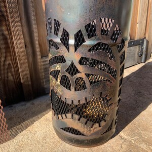 Może przedstawiać: Metalowy kosz na śmieci w kolorze rdzy z wyciętym wzorem tiki. Maska wykonana jest z czarnej metalowej siatki i ma ciemnobrązową patynę.