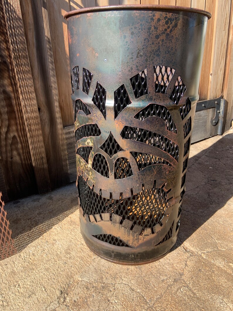 Może przedstawiać: Metalowy kosz na śmieci w kolorze rdzy z wyciętym wzorem tiki. Maska wykonana jest z czarnej metalowej siatki i ma ciemnobrązową patynę.