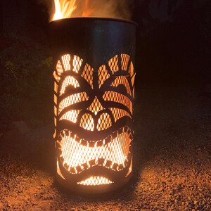 Może przedstawiać: Metalowy palenisko w kształcie maski tiki z płomieniami palącymi się w środku. Palenisko stoi na żwirze.