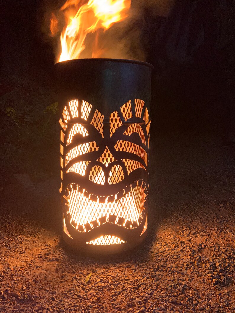Może przedstawiać: Metalowy palenisko w kształcie maski tiki z płomieniami palącymi się w środku. Palenisko stoi na żwirze.
