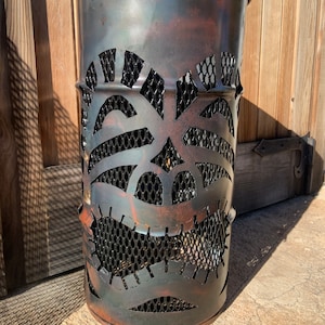 Może przedstawiać: Metalowy kosz na śmieci w kolorze rdzy z wyciętym wzorem twarzy tiki. Kosz ma wewnątrz wyści&oacute;łkę z czarnej siatki drucianej.