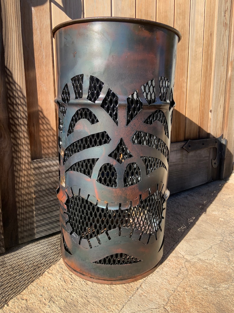 Może przedstawiać: Metalowy kosz na śmieci w kolorze rdzy z wyciętym wzorem twarzy tiki. Kosz ma wewnątrz wyści&oacute;łkę z czarnej siatki drucianej.