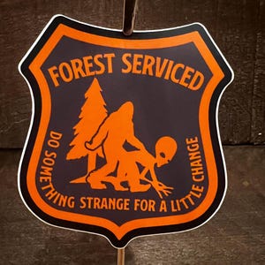 May include: Orange and black sticker with the text "Forest Serviced Do Something Strange For A Little Change" and a silhouette of a Bigfoot and an alien.