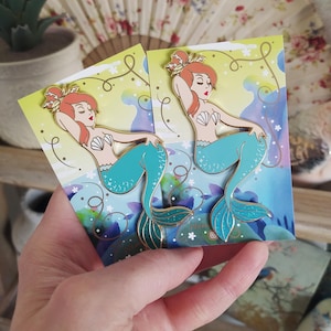 Op de afbeelding: Twee gouden emaillepins met een zeemeermin met rood haar, een schelpbeha en een turquoise staart. De pins zijn geplaatst tegen een achtergrond van witte sterren en blauwe aquarel.