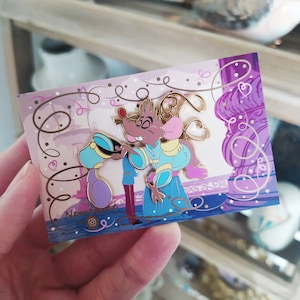 Pode incluir: Um broche de esmalte dourado com um casal de ratinhos de desenho animado em uma pose romântica. O ratinho macho está vestindo um uniforme azul e um chapéu, enquanto a ratinha está vestindo um vestido rosa. O broche está sobre um fundo roxo e dourado com corações e volutas.