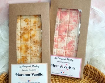 May include: Two brown cardboard boxes with clear windows displaying soy wax melts. The left box features a vanilla scented melt with a light brown color and the text "Macaron Vanille". The right box features a cherry blossom scented melt with a pink and white color and the text "Fleur de cerisier". Both boxes have the text "Les Bougies de Madley" and "Tablette fondante".
