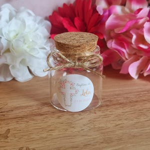 May include: A small, clear glass jar with a cork stopper, tied with twine. The jar has a label with a floral cross design and the text "Baptême de Lola 23.10.2020". The background features white, red, and pink flowers.