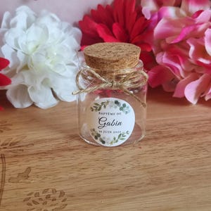 May include: A small, clear glass jar with a cork stopper, tied with twine. The jar has a label that reads "Baptême de Gabin, 16 Juin 2026". The background features white, red, and pink flowers, suggesting a celebratory theme.