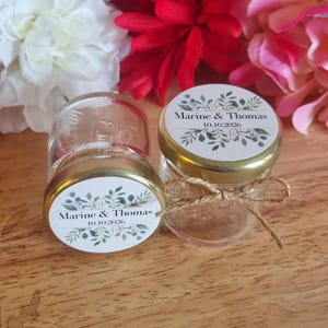 Peut inclure: Deux petits pots en verre avec des couvercles dorés et des étiquettes indiquant "Marine & Thomas" et "10.10.2026". Chaque pot est noué avec de la ficelle. Le fond comprend des fleurs blanches, rouges et roses.