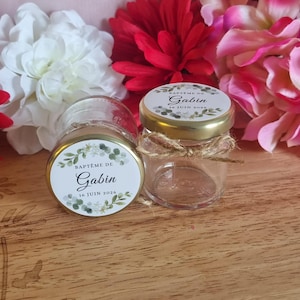 Può includere: Due piccoli barattoli di vetro con coperchi dorati ed etichette decorative. Le etichette recitano "Baptême de Gabin, 16 Juin 2026" e presentano un motivo floreale. Un barattolo ha un fiocco di spago. I barattoli sono su una superficie di legno, circondati da fiori.