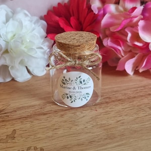 Può includere: Un piccolo barattolo di vetro trasparente con un tappo di sughero, legato con spago. Un'etichetta rotonda sul barattolo recita "Marine & Thomas" e "10.10.2026" circondata da un disegno floreale. Il barattolo è su una superficie di legno, con fiori rosa e rossi sullo sfondo.