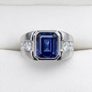 May include: A silver-toned ring featuring a large, rectangular, emerald-cut blue sapphire at the center. The band has a brushed metal finish and is accented with two small, round, clear stones on each side of the central sapphire setting.