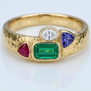 Op de afbeelding: Een gouden ring met een gehamerde textuur, voorzien van een centrale smaragdgeslepen groene edelsteen, een kleine ovale witte steen, een driehoekige rode steen en een driehoekige blauwe steen in een clusteropstelling.