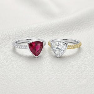 May include: Two rings featuring trillion-cut center stones. The left ring has a deep red ruby set in a silver-toned band with pavé diamonds. The right ring has a clear, faceted stone set in a gold-toned band with pavé diamonds along the shank.