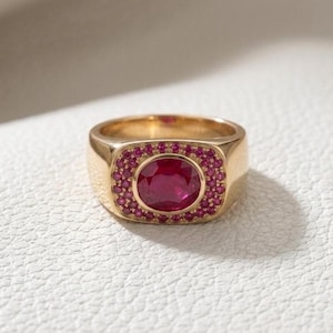 May include: A gold ring featuring a large, oval-cut central red gemstone surrounded by a halo of smaller, round-cut red stones set into a rectangular frame. The band is smooth and polished, showcasing a classic design with vibrant red accents.