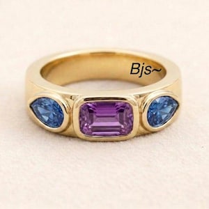 Op de afbeelding: Een gouden ring met een centrale rechthoekige paarse edelsteen, geflankeerd door twee peervormige blauwe edelstenen. De ring heeft een gladde, gepolijste afwerking en de tekst Bjs~ is zichtbaar aan de binnenzijde van de gouden ringband.