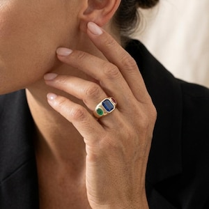 Peut inclure: Une bague en or ornée de trois pierres précieuses colorées : verte, bleue et rouge. La bague a une forme rectangulaire avec un anneau en or poli. Les pierres sont disposées en rang, créant un design audacieux. La bague est portée au doigt.