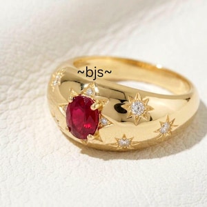 Op de afbeelding: Een gouden ring met een centrale ovaal geslepen rode edelsteen, omringd door stervormige gravures die elk zijn bezet met een kleine heldere diamant. De ring heeft een gladde, gepolijste afwerking en bevat de tekst ~bjs~ op het bovenoppervlak.