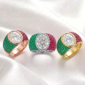 Peut inclure: Trois bagues ornées de pierres précieuses avec une pierre centrale de forme ovale. Les bagues présentent une combinaison de pierres vertes et rouges, avec un anneau en or, argent et or rose. Les bagues sont posées sur un fond blanc.