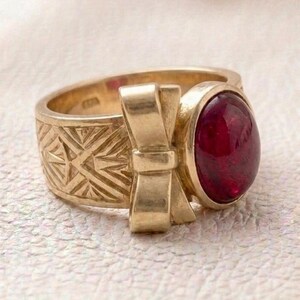 Könnte beinhalten: Ein goldener Ring mit einem polierten, tiefroten ovalen Cabochon-Edelstein in einer Fassung. Das Band ist mit einem erhabenen, dreidimensionalen Schleifenmotiv und komplizierten geometrischen, fächerartigen Gravuren auf der Metalloberfläche verziert.