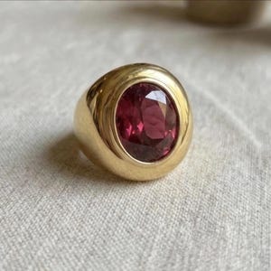 Könnte beinhalten: Ein Goldring mit einer glatten, polierten, breiten Ringschiene und einem markanten, oval geschliffenen, tiefrosa Edelstein in der Mitte. Das Metall hat eine reflektierende Hochglanzoberfläche, die einen Kontrast zum dunklen, durchscheinenden Stein bildet.