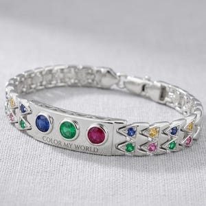 Op de afbeelding: Zilveren armband met een reeks kleurrijke edelstenen. De armband heeft een rechthoekig middelpunt met de tekst "COLOR MY WORLD" en drie ronde edelstenen. De armband is versierd met diverse driehoekige en ronde edelstenen.